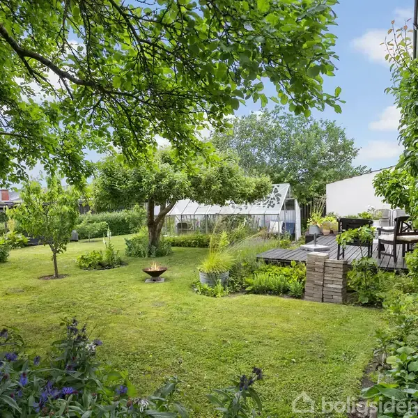 En grøn have med en bålplads og træer. En terrasse med havemøbler ligger til højre, omgivet af blomster og buske. I baggrunden ses et drivhus.