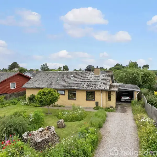 Et gult murstenshus med fladt tag står omgivet af en grøn have. Der er en grusvej og carport ved siden af huset. Omkring ses andre parcelhuse.