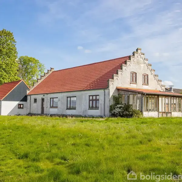 Et hvidt hus med rødt tegltag står stille på en grøn græsplæne, omgivet af træer og en blå himmel.