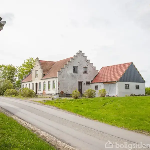 Et gammelt landhus med hvidmalet facade og rødt tegltag står langs en smal vej. Omkring er der grønne plæner og træer. Ingen tekst er synlig.