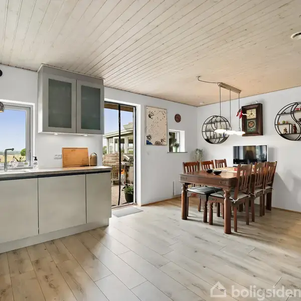 Et moderne køkken med hvide skabe ved siden af et spisebord med træstole. Rummet er lyst og åbent med store vinduer, og der er en dør til en terrasse.