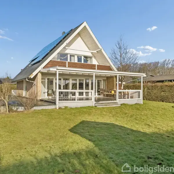 Et moderne hus med skråt tag og solpaneler står på en velplejet græsplæne. En overdækket terrasse rummer udendørs møbler. Buske og træer omkranser haven.