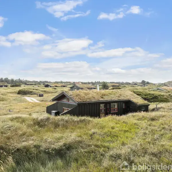 Et sort, træhus ligger stille i en åben mark af højt græs under en klar blå himmel med lette skyer. Bankerne af jord og græs starter i horisonten.