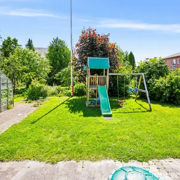 Lille legeplads med legetårn og rutsjebane står på en græsplæne. I baggrunden ses en række buske, et drivhus og et hus. Klar himmel og sollys.