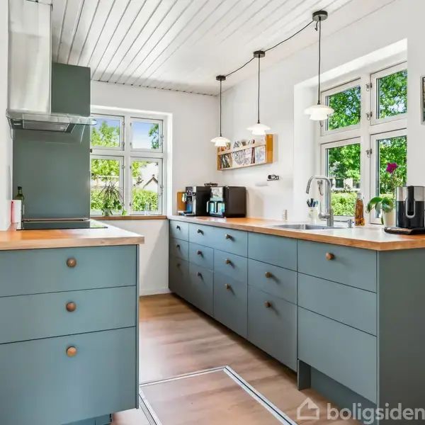 Køkken med grønne skabe og træbordplader, arrangeret med kaffemaskiner og en brødrister. Store vinduer bagved slipper naturligt lys ind, og to vedhængslamper hænger over bordpladen.