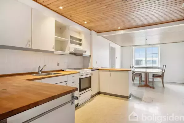 Køkken med hvide skabe og træbordplader står i et lyst rum. I baggrunden ses et spisebord ved et vindue. Loftet er beklædt med træpaneler.