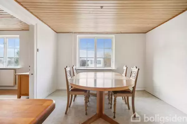 Et ovalt træbord med seks stole står stille i et lyst rum med træloft og hvide vægge. Et vindue i baggrunden slipper dagslys ind.