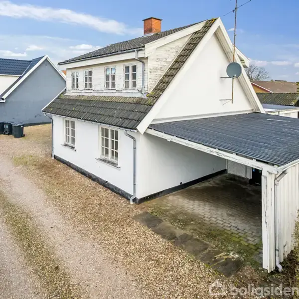 Et hvidt hus med to etager har et sort tag og en tilstødende carport. Det står ved en grusvej, hvor der ses flere omkringliggende huse og grønne træer i baggrunden.