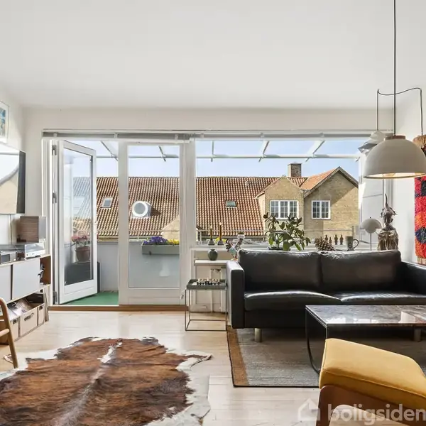 Stue med sort lædersofa, åbne skydedøre til balkon; stuebord og lammeskindstæppe på gulvet. Væggene prydes af kunst, hylder og lamper. Udsigt til bygninger med røde tage.