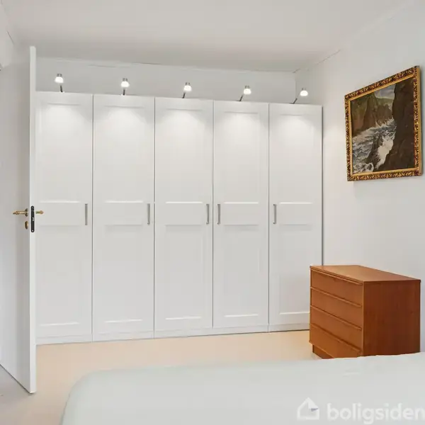 Et hvidt garderobeskab står oplyst mod en væg i et soveværelse med beige gulv. Ved siden af er en brun kommode, og kunst hænger på væggene.