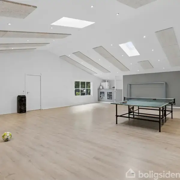 Et stort rum med trægulv rummer et bordtennisbord og en gul fodbold, mens et sammenklappeligt rebspil står i hjørnet. Loftet har ovenlys, der giver lysindfald.