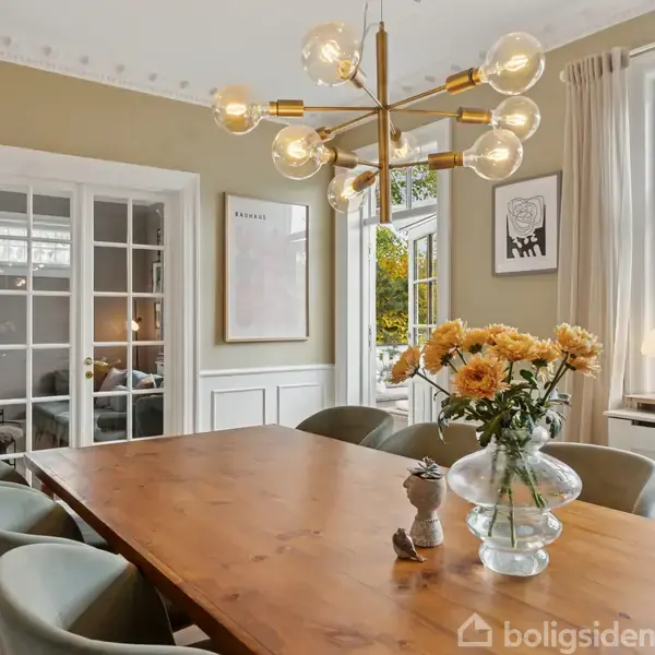 En spisestue med et træbord dekoreret med en vase med blomster. Moderne lysekrone hænger over bordet. Store vinduer slipper naturligt lys ind i det elegante interiør. En plakat med teksten "SABANÅS" hænger på væggen.