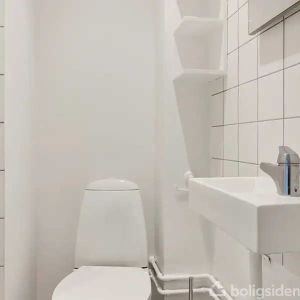 Toilet uden låg står ved siden af en lille håndvask med moderne vandhane. Flisebelagte vægge danner et hvidt, minimalistisk badeværelsesmiljø.