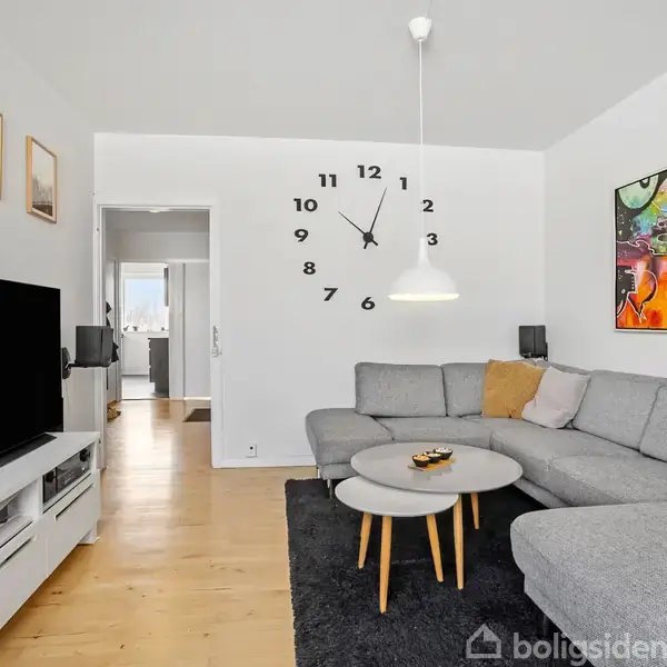 Gråt hjørnesofa står mod hvid væg med stor ur. Lige foran er et rundt sofabord på sort tæppe. Et stort fladskærms-tv er placeret til venstre. Vægkunst hænger oppe.