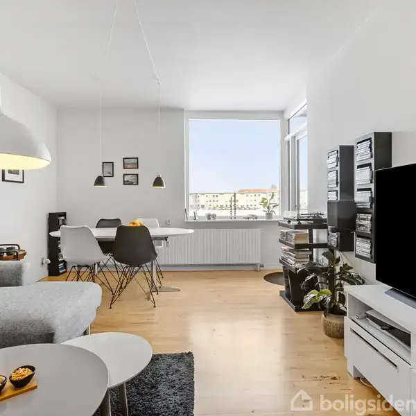 En lys stue med gråt sofaarrangement med orange pude, rundt sofabord og skål med snacks. Fladskærms-tv på hvid reol. Spisebord med fire stole ved vindue og udsigt.