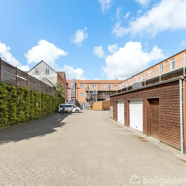 En parkeringsplads med en bil holder stille. Til venstre er et hegn med grønt løv, og til højre står røde garager op ad bygninger under en blå himmel.