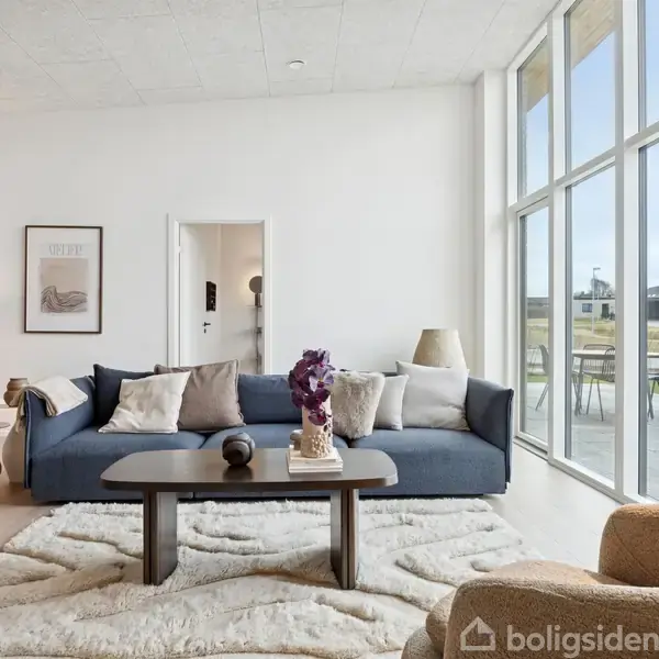 En stue med en blå sofa placeret på en lys, fluffy gulvtæppe. Et sofabord står foran sofaen, dekoreret med en vase og bøger. Store vinduer giver udsigt til en terrasse og have.