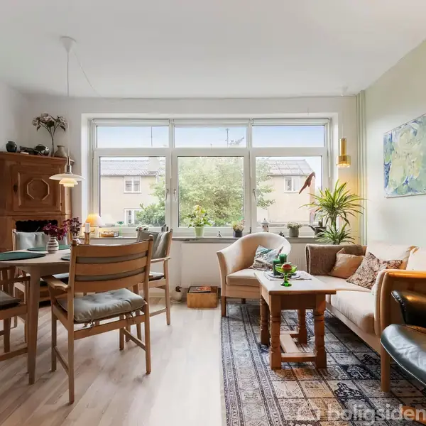 Stue med spisebord og stole; sofaer og lænestol omkring sofabord; vindue med udsigt til træer og bygninger udenfor; dekoreret med planter og kunstværker på væggen.