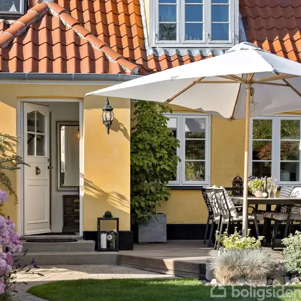 Et gult hus med rødt tegltag har en terrasse med et spisebord og stole under en stor parasol. Omkring terrassen er der blomster og grønne planter.