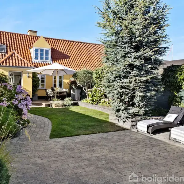 En gårdhave med terrassesten, to liggestole, en stor grantræ, blomsterbede og en gul villa i baggrunden. Et hvidt parasol står over et bord og stole.