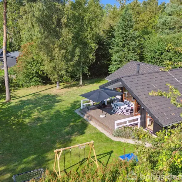 Et træhus med terrasse, hvor der står et bord og stole under en parasol. Huset er omgivet af en frodig have med græsplæne og store træer.