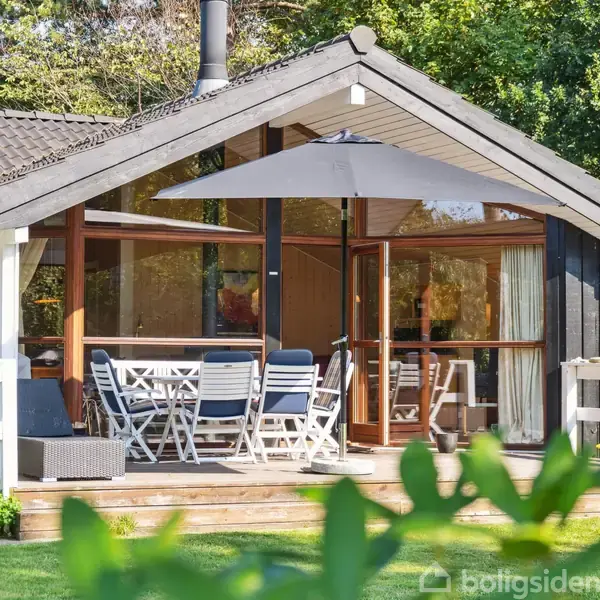 Et sort træsommerhus med store vinduer har en terrasse med udendørs spisebord og parasol. Det står omgivet af grønne træer og en velplejet græsplæne.