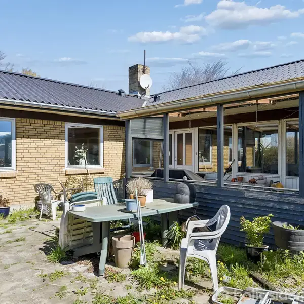 Et udendørsområde med havemøbler står på en flisebelagt terrasse foran et murstenshus. Omkring terrassen ses blomsterpotter og planter, og huset har store vinduer og et enkelt skorstensrør.