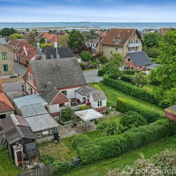 Huse med røde tage står tæt i en landsby, omringet af grønne haver og træer. I baggrunden ses havet under en blå himmel.