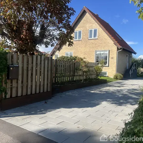 Et gult murstenshus med rødt tag står i sollys. En flisebelagt indkørsel fører til hoveddøren. Træhegn omkranser forhaven med grønne planter. Husnummeret 28 ses tydeligt.