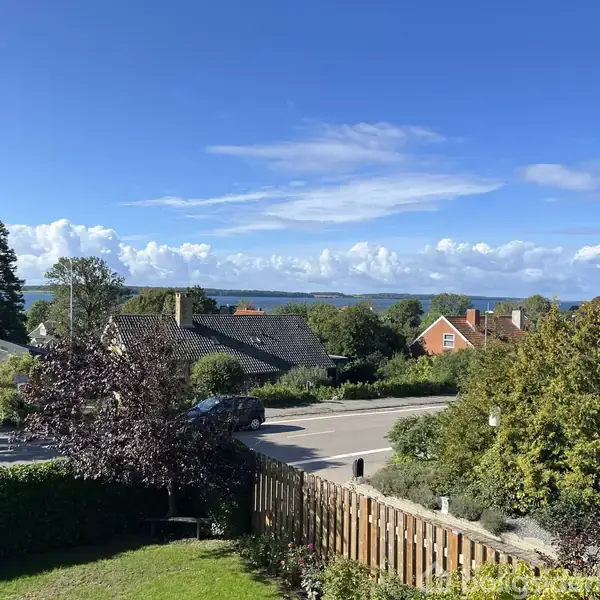 Huse står stille på en bakke med en udsigt over træer og en fjern horisont med havudsigt. Skyer og blå himmel skaber en lys og fredelig atmosfære.