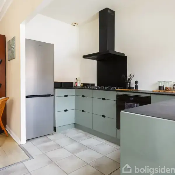 Gråt køleskab står i et moderne køkkenhjørne med grønne skabe og sort emhætte. Ved siden af er en træstol. I baggrunden ses en gang og et værelse.