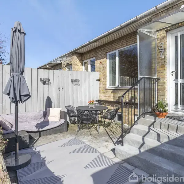 Husets indgangsparti består af en trappe, der leder op til en hvid dør. En terrasse med grå parasol, sofa og rund bordgruppe står i gårdhaven. Huset er murstensbygget. Nummer 37 vises ved døren.