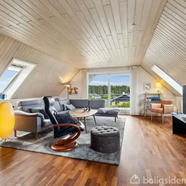 En stue med skrå lofter har en sofa og lænestol på et tæppe, med en gul lampe til venstre. Et stort vindue viser træer udenfor. En fladskærm er til højre.