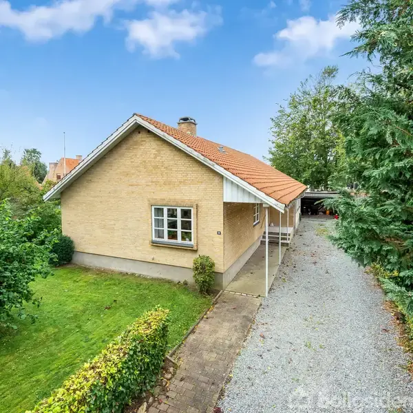 Et enkelt hus med rødt tag står som hovedfokus; det står i en grøn have med græsplæne og buske. Til højre løber en grusbelagt indkørsel ved siden af.