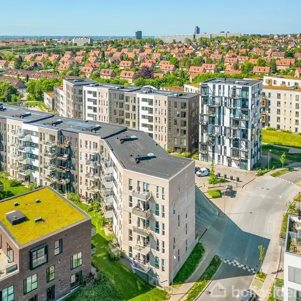 Moderne lejlighedsbygninger står stille i et urbaniseret område med velplejede grønne tage, omgivet af veje og grønt areal. I baggrunden ses en by med ensartede, røde rækkehuse.
