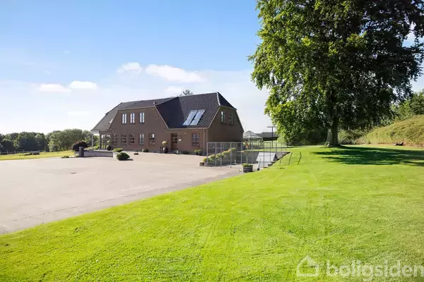 En stor murstensbygning står på en åben plads omgivet af grønne græsplæner og et stort træ. Bygningen har flere vinduer og et mørkt tag.