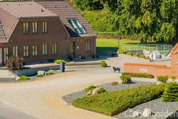 En sort hund står på en grusindkørsel foran et stort murstenshus med rødt tag. Omkring er der trimmede buske og et grønt område med bure. Skilt med "Risdal".