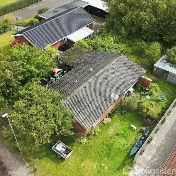 Et hus med et sort tag er omgivet af en grøn have. Der står en bil i indkørslen, og træer omgiver ejendommen. Der er en sti ved siden af huset.