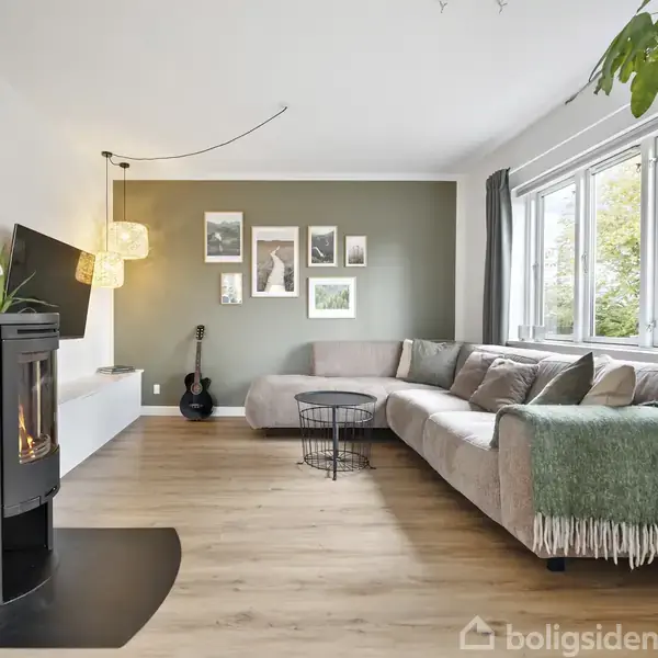 En moderne stue med en brændeovn i brug. En stor beige sofa står ved vinduet, dekoreret med puder og et grønt tæppe. Kunstværker og planter pryder rummet.