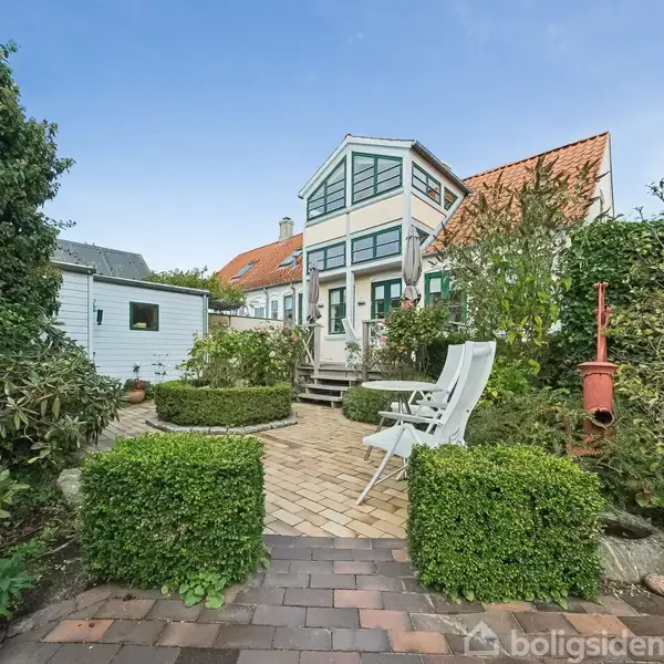 Et hvidt hus med mange vinduer står i en velplejet have med brostensbelægning. To hvide havestole står foran huset omgivet af grønne buske. En rød postkasse er placeret til højre.