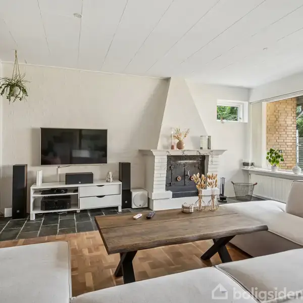 Tv på hvidt møbel står i lys stue ved siden af pejs, omgivet af brun sofa og træbord, med vindue mod terrasse med planter og udsigt til have.