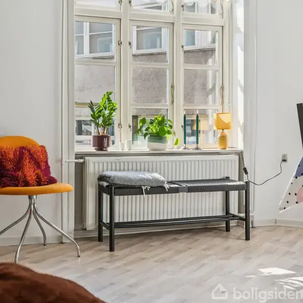 En orange stol med pude står ved et stort vindue med planter og lampe, ved siden af en bænk. I nærheden er et spisebord dækket med en prikket dug.