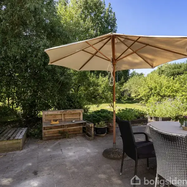 Parasollen står åben over et bord med stole på en flisebelagt terrasse. Omgivet af grønne træer og buske, skaber det en hyggelig udendørs atmosfære.