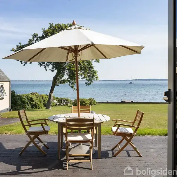Et rundt træbord med fire stole står under en parasol på en terrasse. I baggrunden ses en græsplæne, træer, og havudsigt med en båd i horisonten.