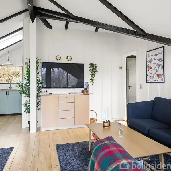Stue med mørkeblåt sofaarrangement foran et sofabord, grene og planter hænger fra væggen. Køkken i baggrunden med lyseblå skabe. Loft har synlige sorte bjælker og ovenlys.