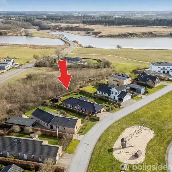 Hus markeret med rød pil i boligområde. Flere huse omkring, tæt på en vej med grønne arealer. En flod og skov ses i baggrunden.