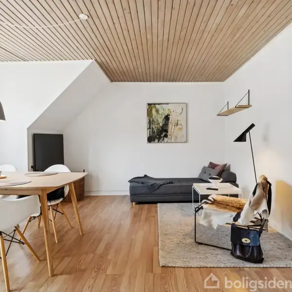 Stue med træloft og trægulv indeholder et spisebord med hvide stole til venstre og en grå sofa til højre. Et sofabord står på et tæppe ved siden af en gulvlampe.