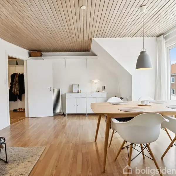 Et lyst spisestueområde med et træbord og fire hvide stole. Bordet er placeret nær et stort vindue, der giver naturligt lys. Loftet er træbeklædt, og der hænger en sort pendellampe over bordet.