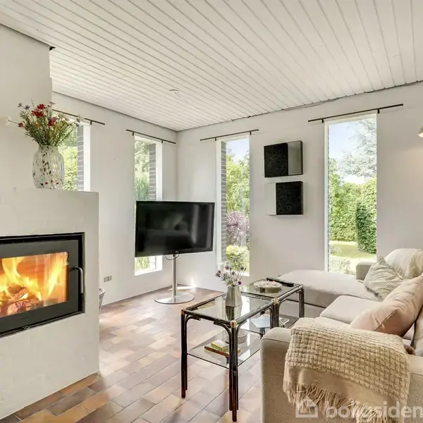 Brændeovn brænder lystigt i en stue med en beige sofa. Glasbord foran sofaen. Store vinduer lader naturligt lys ind, og der er en vase med blomster på ovnen.