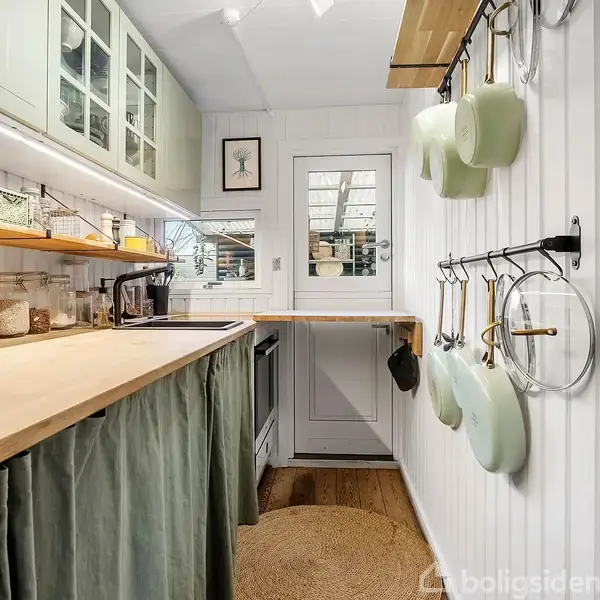 Et lille køkken med træbordplader og gryder hængende på væggen. Skabe og hylder opbevarer redskaber, mens et tæppe ligger på det trægulv. En dør fører ud til et andet område.